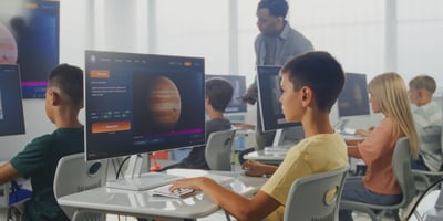 Students in a classroom use desktop computers to explore an interactive space simulation while a teacher assists them.