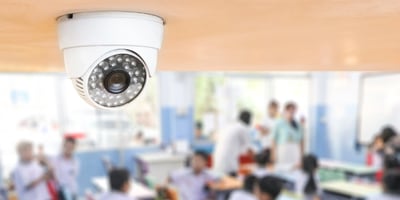 A white dome-style security camera mounted on a ceiling monitors a bright classroom, with students seated at desks and teachers standing and interacting in the softly blurred background.