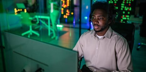 A person wearing a headset works at a computer in a modern office illuminated by green and blue lighting.