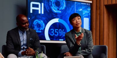 Two individuals in professional attire discussing something in front of them with a large display in the background.