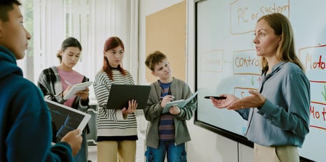 Woman in professional attire teaching four kids on tablets in front of a large display.