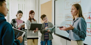 Woman in professional attire teaching four kids on tablets in front of a large display.