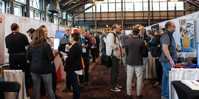 Room of people exploring an exhibit hall lined with vendors in pipe-and-drape booths.