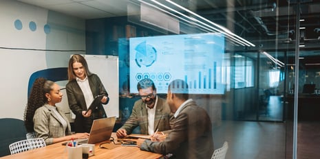 Four professionals collaborate in a modern glass-walled meeting room, reviewing data on a large digital display while discussing insights around a table with laptops and tablets.
