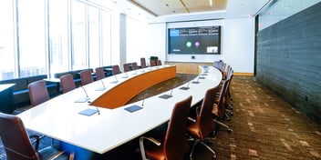 Large boardroom with many chairs arranged in an oblong pattern facing a large monitor.