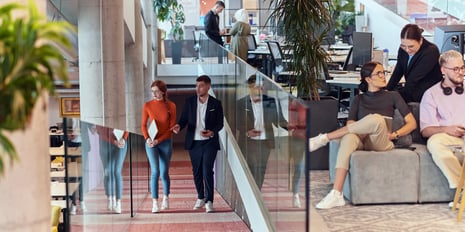 In a modern open office, a woman in an orange sweater and jeans walks with a man in a black suit, while nearby colleagues in casual and business attire collaborate and relax amid greenery and sleek furniture.