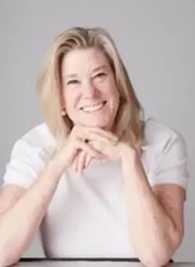 A smiling woman with shoulder-length blonde hair sits with her hands clasped in front of her, wearing a simple white short-sleeved shirt and gold hoop earrings against a plain gray background.
