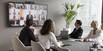 Four people dressed in business professional taking part in a hybrid meeting looking at a large display with others joining virtually.