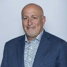 A bald, middle-aged man in a blue blazer and patterned shirt smiles slightly while posing against a plain light background.