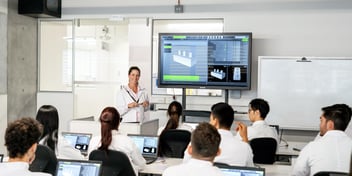 woman standing in front of a class of students teaching from a large display