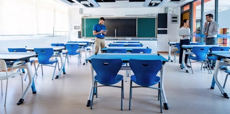 Bright blue chairs and white desks fill a modern classroom while three educators prepare and discuss plans near the chalkboard and windows.