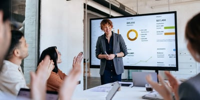 A smiling presenter stands beside a large screen showing financial charts and metrics while colleagues seated around a table applaud during a business meeting.