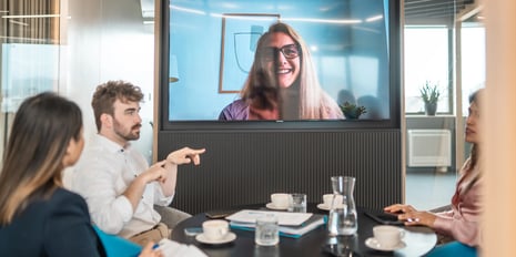 Three young individuals taking part in a hybrid meeting with one participant joining via the large display in front of them.