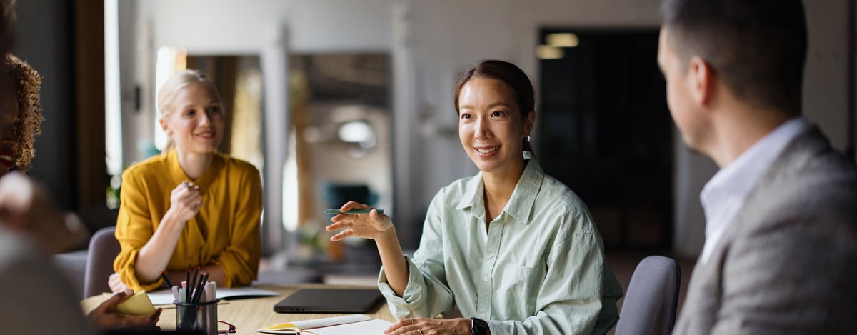 Four professionals sit around a wooden conference table in a modern office as a woman in a light green shirt speaks and gestures with a pen while her colleagues listen and smile.