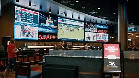 People standing around a casino bar area under a large LED display.