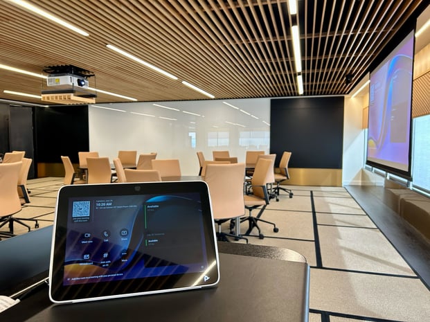 A modern, empty conference room with rolling chairs, a large presentation screen, a ceiling-mounted projector, and a touchscreen meeting control panel in the foreground.