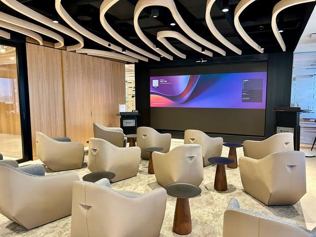 A modern presentation lounge with curved ceiling panels, cushioned lounge chairs, and a large wall-mounted digital display.
