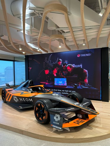An orange-and-black Formula E race car displayed indoors in front of a large digital screen.