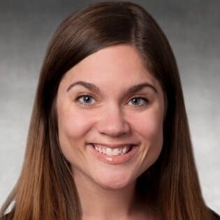 Professional headshot of a person with long brown hair facing the camera against a neutral gray background.
