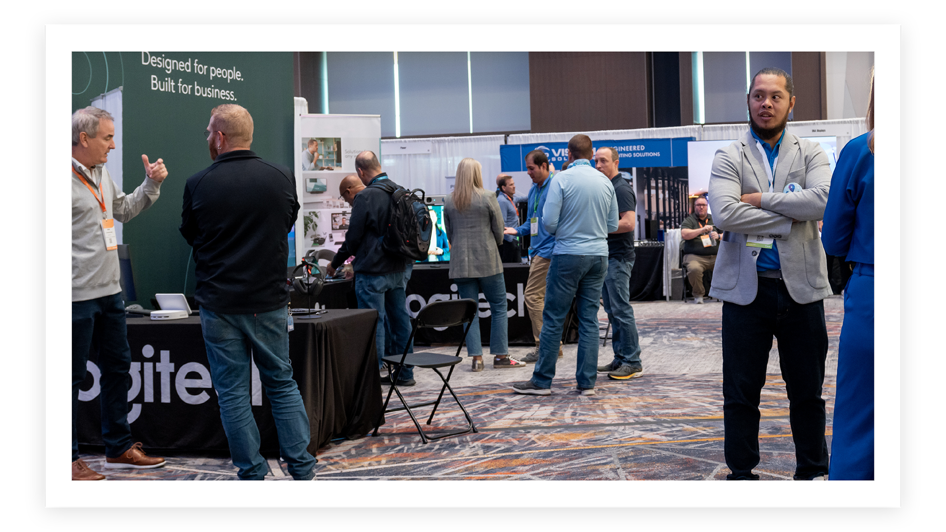 A group of people wearing conference badges stand and talk at exhibitor booths in a carpeted convention hall, with branded displays and product tables visible in the background.