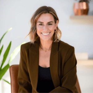 Professional headshot of a person seated indoors, wearing a dark blazer and black top, with a softly blurred background featuring a plant and home office décor.