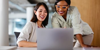 Two professional women smiling and collaborating while looking at a laptop together in a modern office.