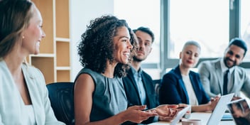 Five people lined up in a boardroom setting while a professionally dressed woman speaks to the group.