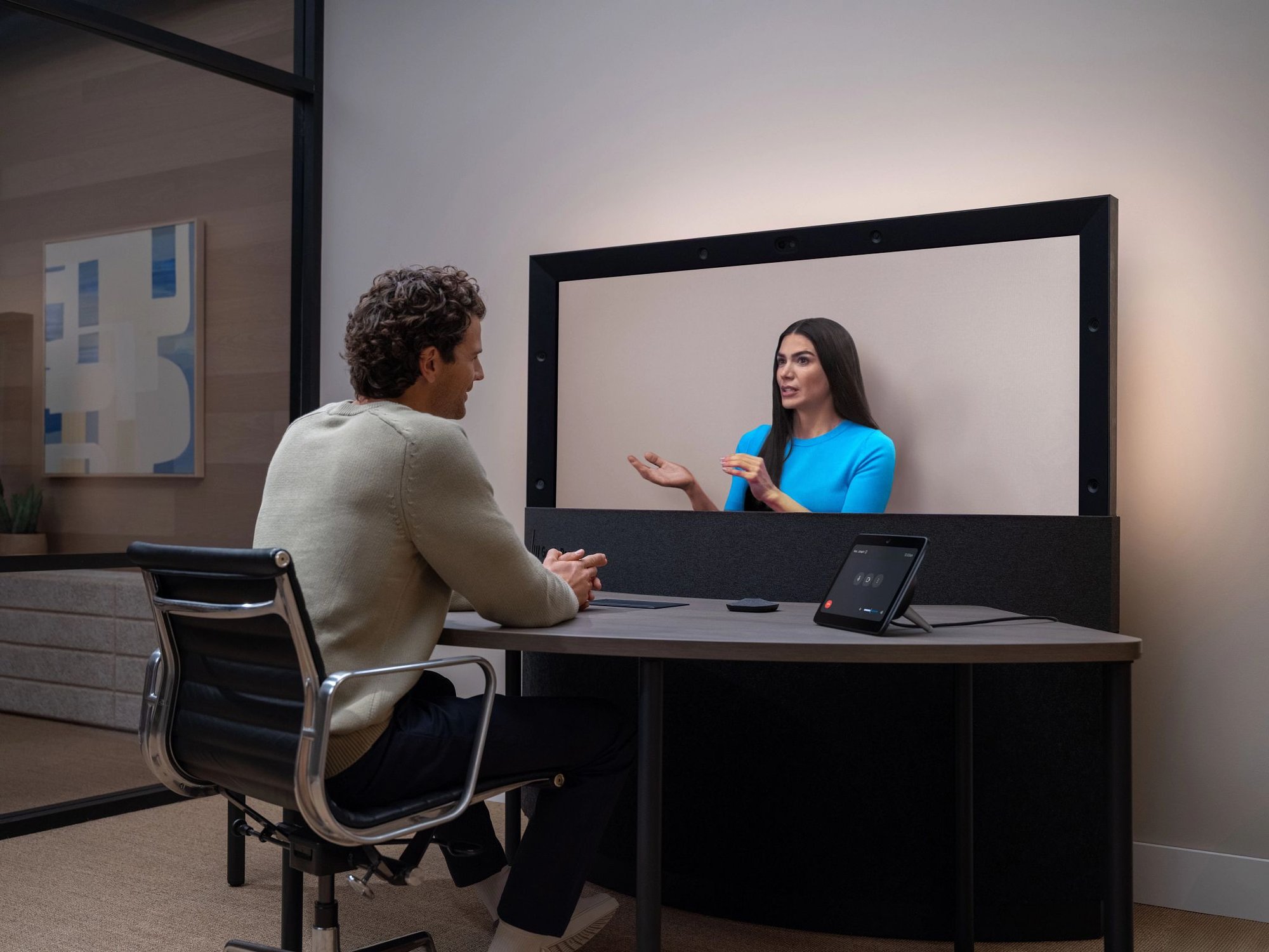 A person seated at a desk engages in a lifelike video meeting with a remote participant displayed on a large Google Beam screen, showcasing an immersive, face-to-face conferencing experience.
