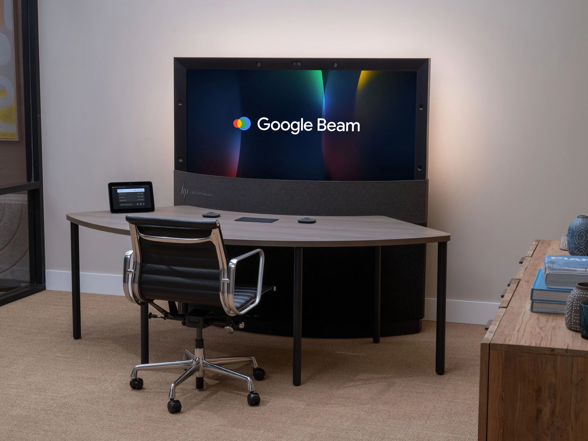 A modern conference room setup featuring a Google Beam video display mounted above a curved desk with a touchscreen controller and chair, designed for immersive virtual meetings.