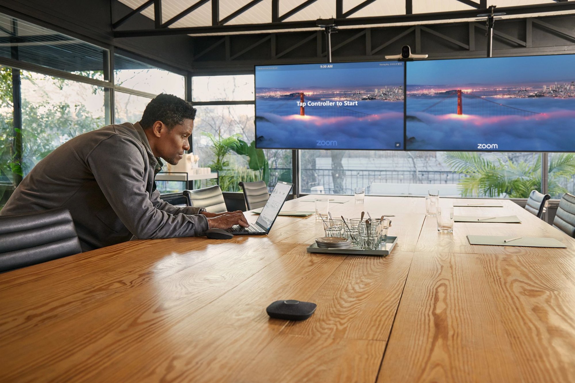 A man sits at a large wooden conference table using a laptop in a modern meeting room, with dual wall-mounted screens displaying a Zoom meeting setup in the background.