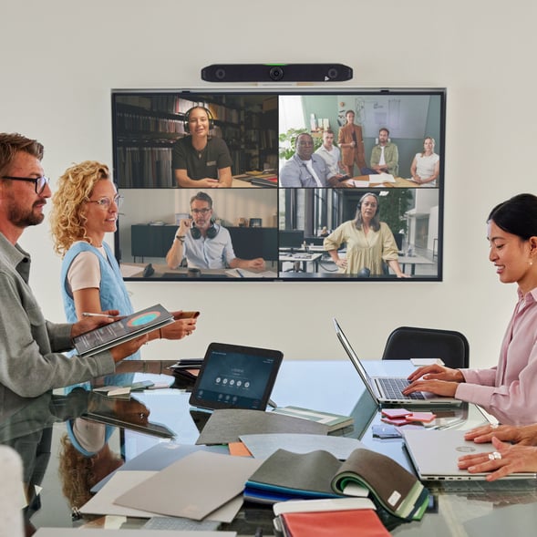 A group of coworkers collaborates around a conference table while joining a video call on a large screen showing multiple remote participants, with laptops, tablets, and meeting materials spread out in a modern office.