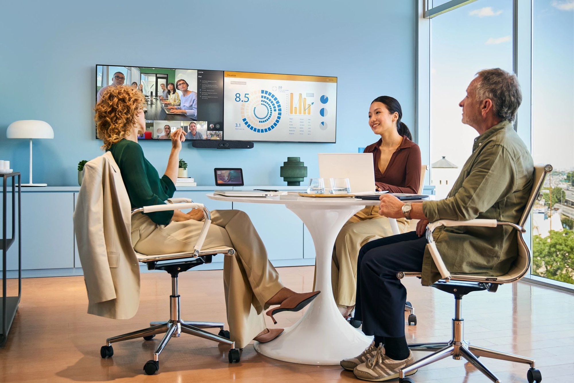 Four colleagues sit around a small table in a modern office, discussing data displayed on a large screen during a hybrid meeting with remote participants visible on a video call.