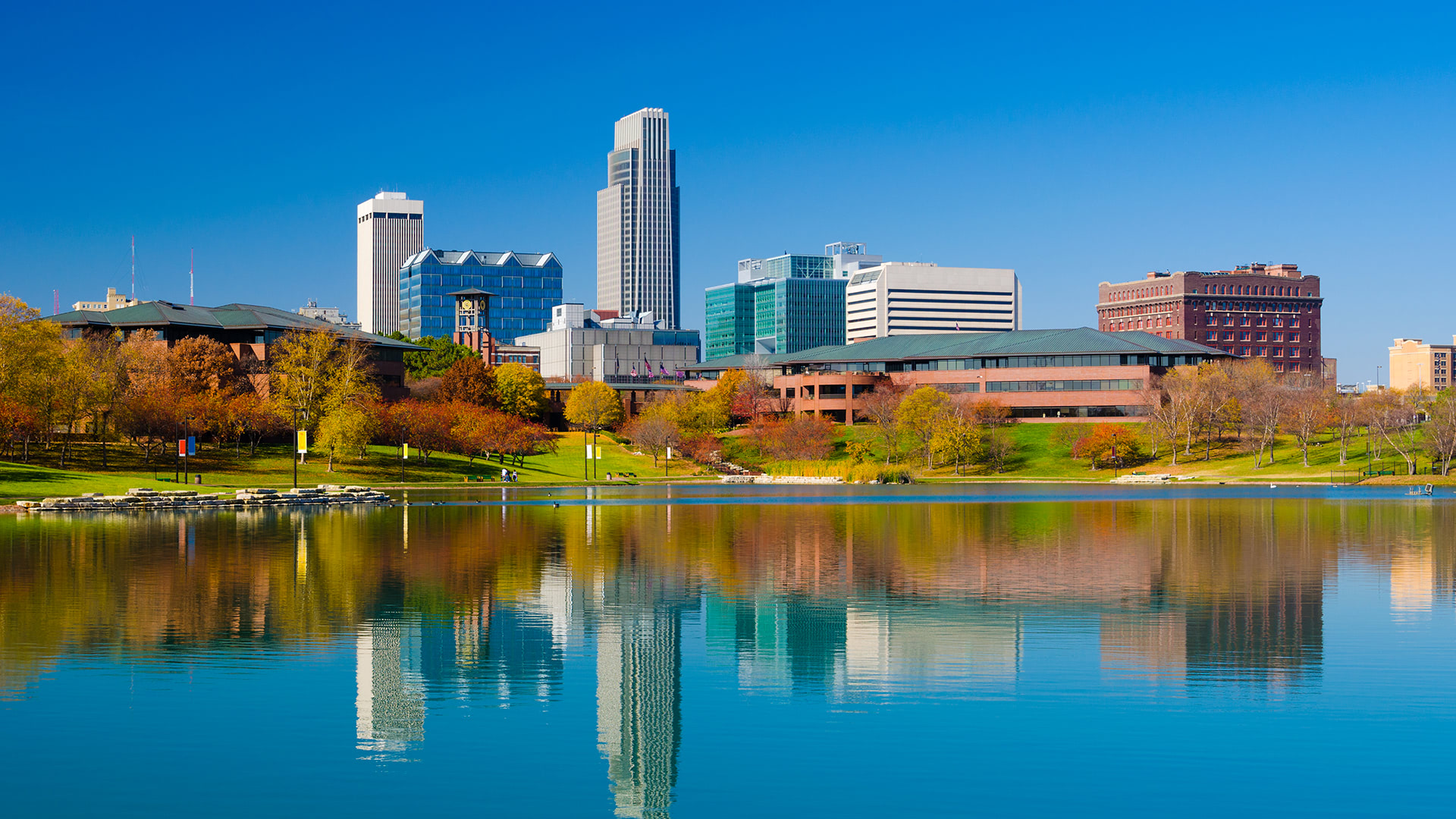 Omaha skyline reflected in a lake on a clear day, showcasing diverse architecture and greenery.