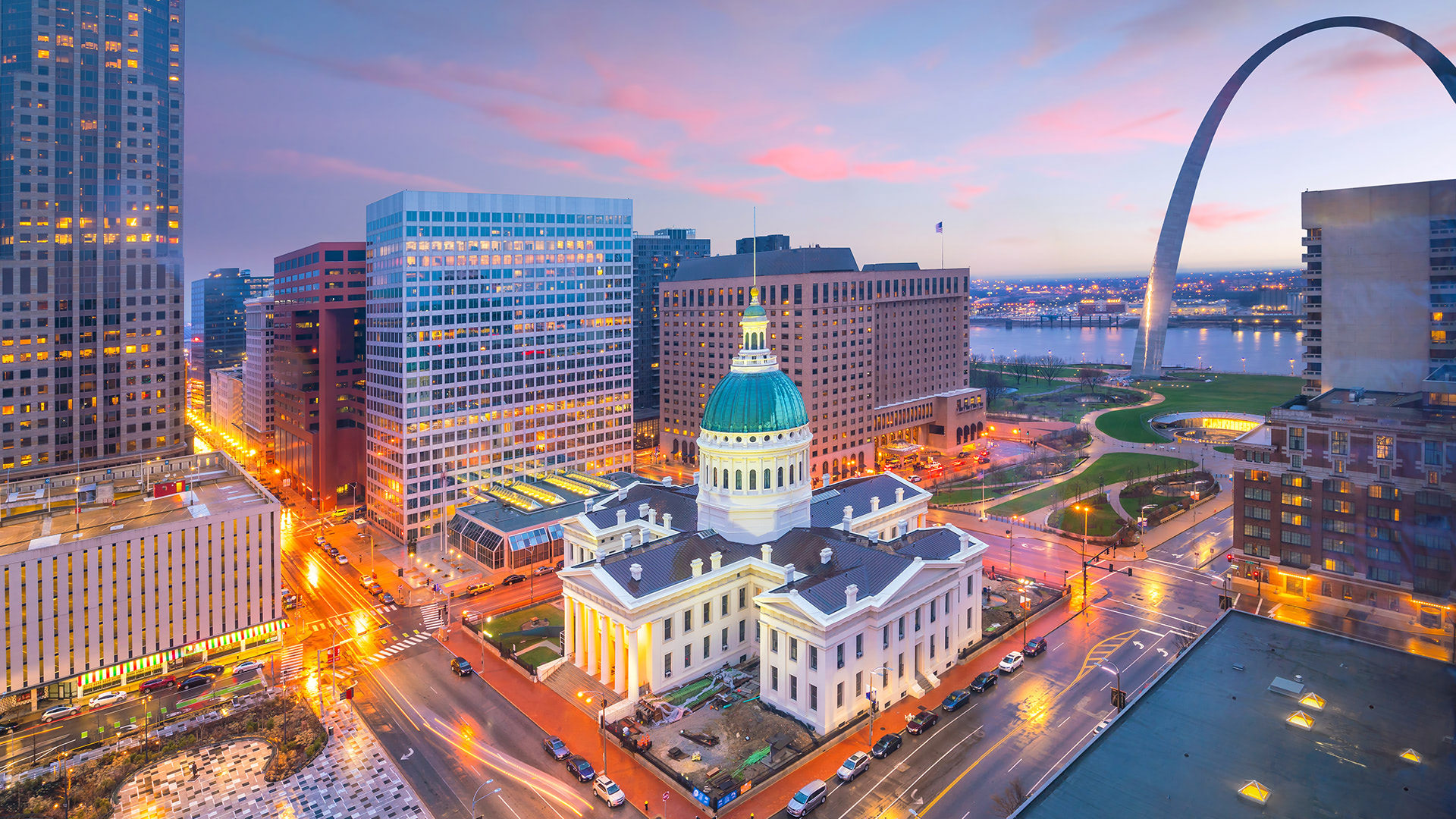 Aerial view of the St. Louis skyline, featuring the Old Courthouse and the Gateway Arch, under a colorful evening sky.