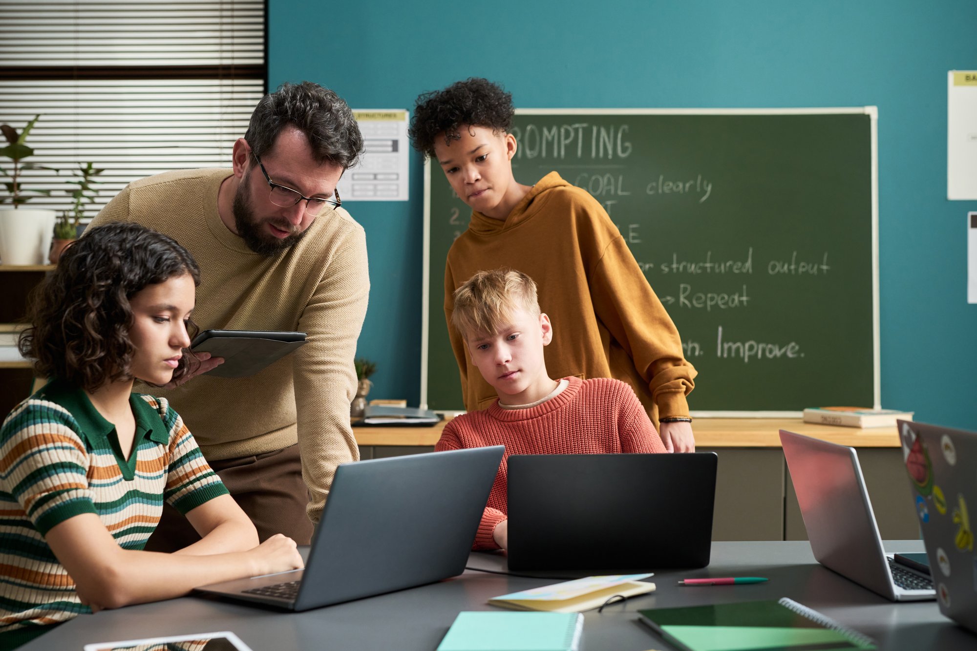 In a bright classroom, a teacher assists three students—one in a striped shirt, one in a hoodie, and one in a sweater—working on laptops, with a chalkboard displaying 