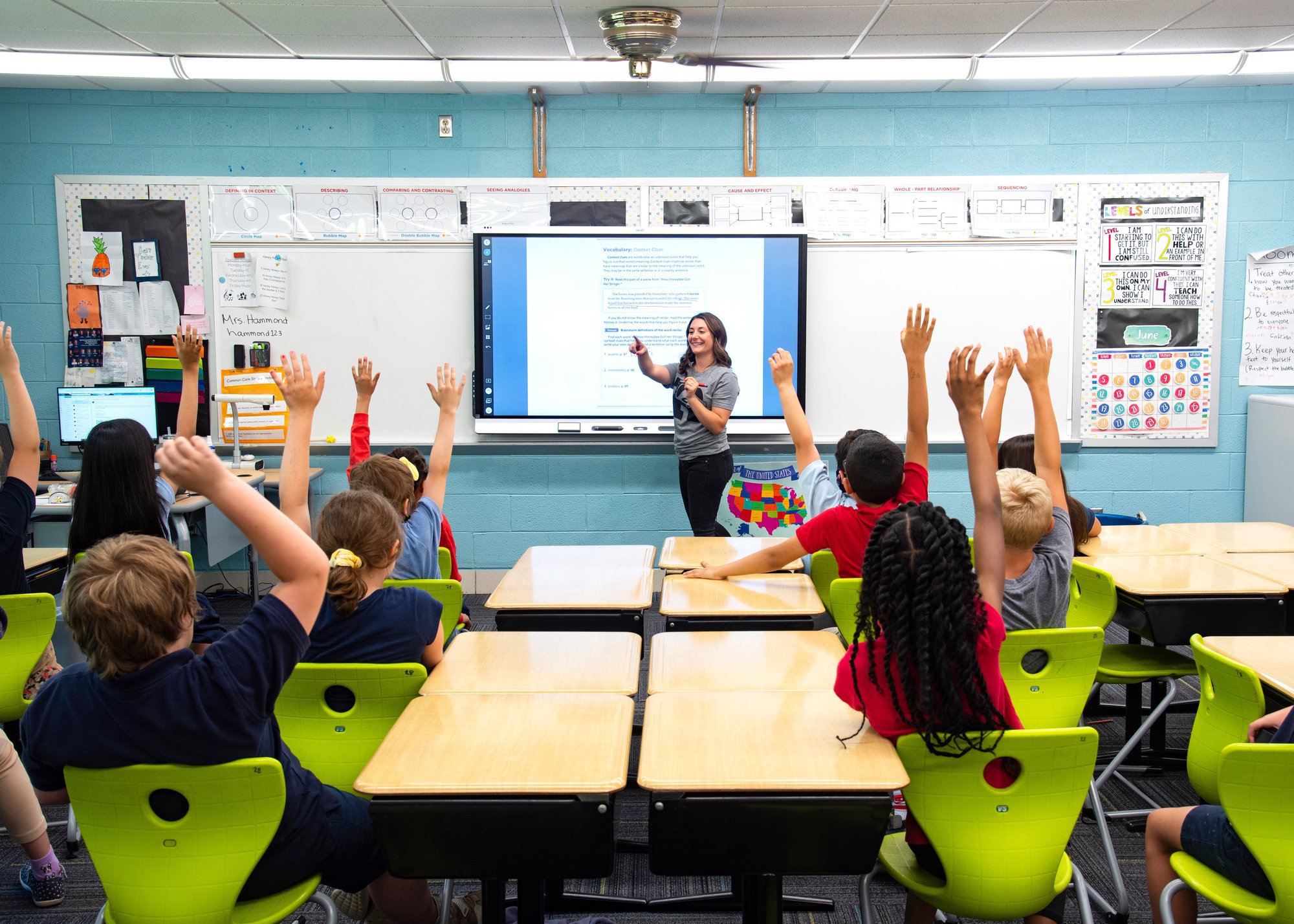 In a brightly lit classroom with turquoise walls and lime green chairs, a smiling teacher stands before an interactive whiteboard, engaging a group of attentive students who enthusiastically raise their hands to answer her question.